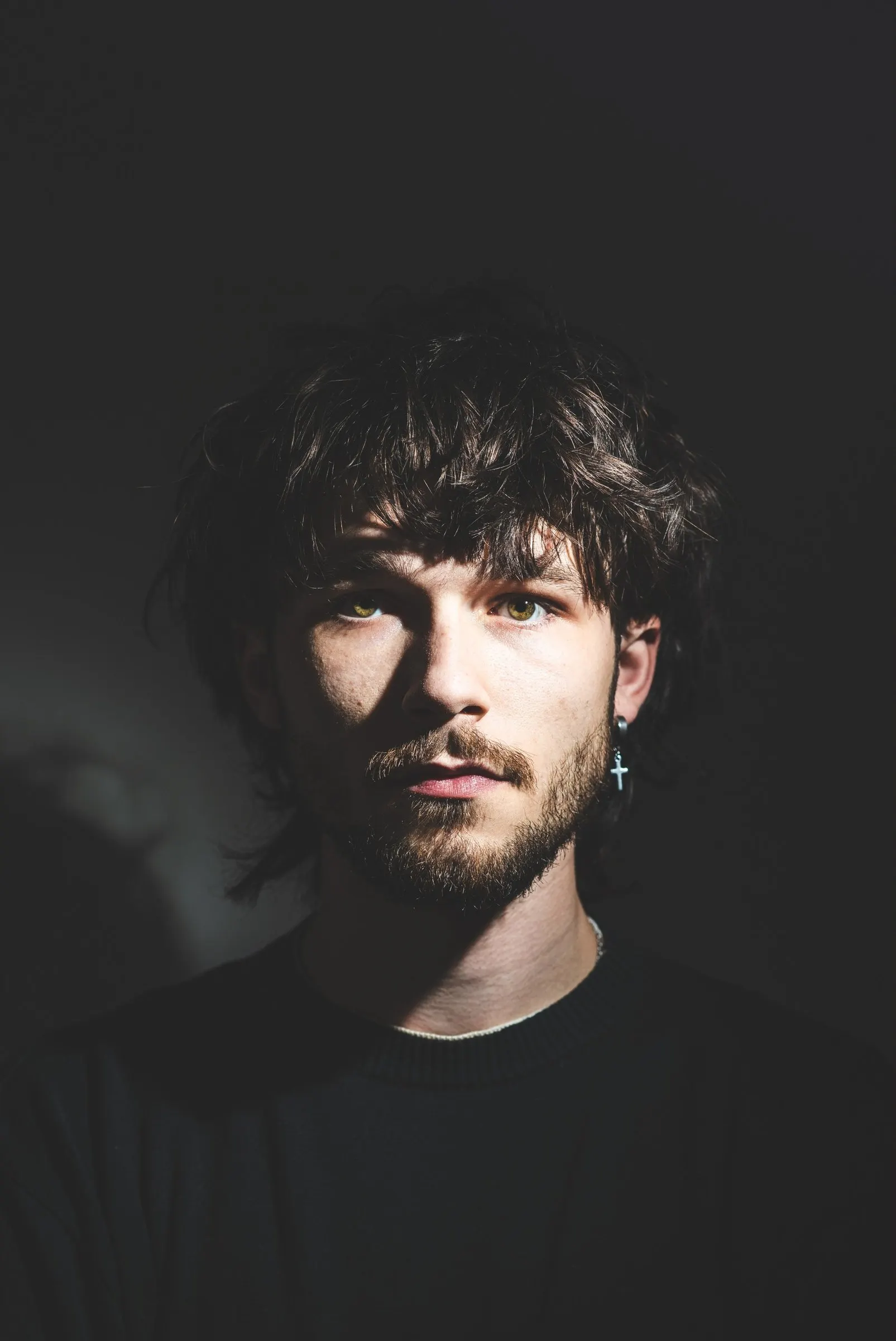 Portrait of Joey, photographer based in Haaksbergen: dark tousled hair, a beard and a cross earring, with hard light across his face against a dark background