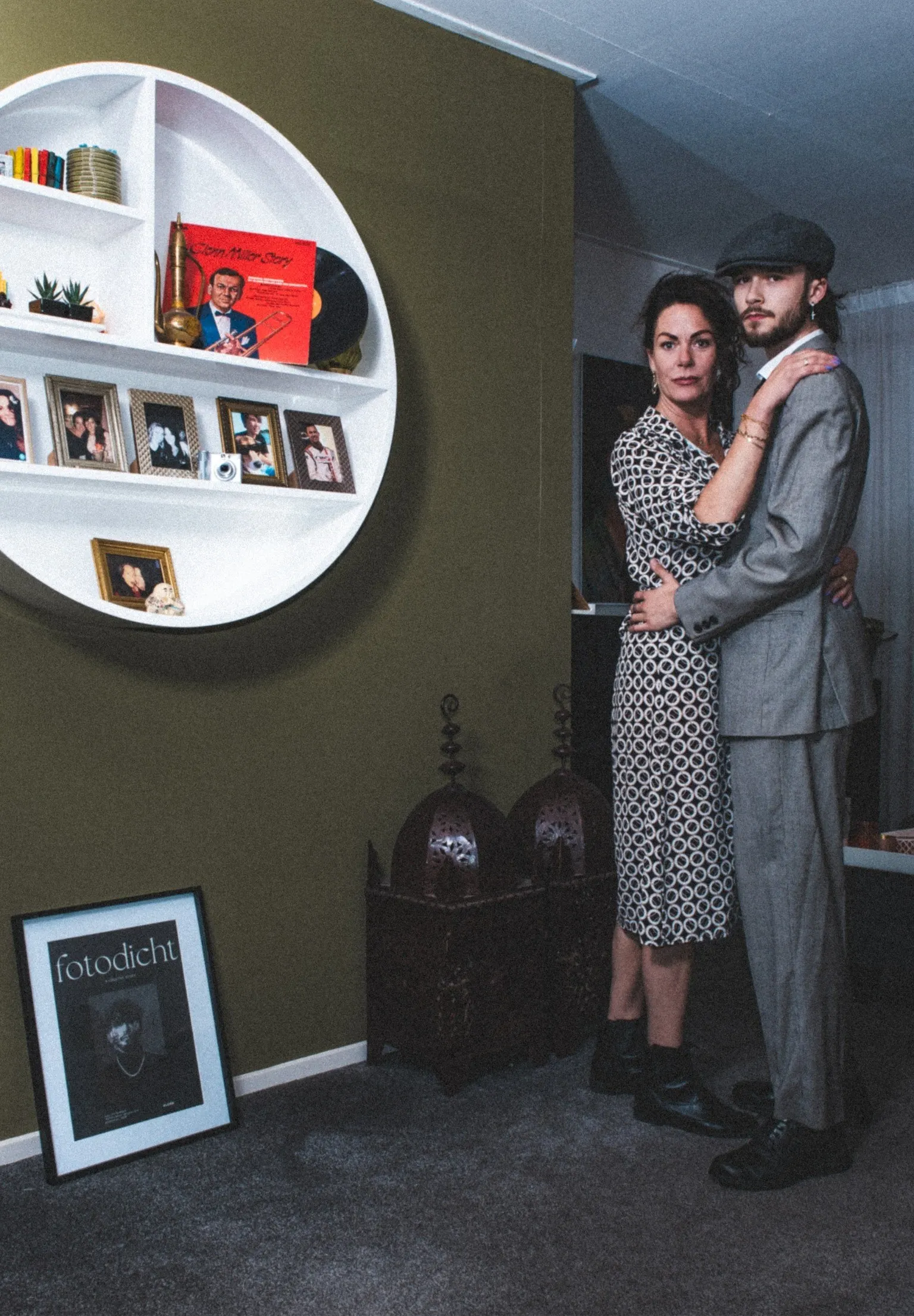 Couple in a living room beside a round wall shelf with records and photo frames; she in a black and white dress, he in a grey suit and cap