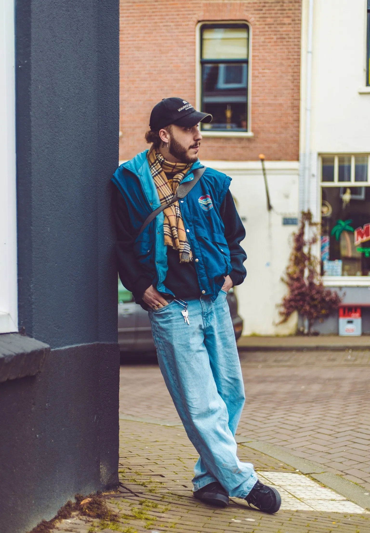 Young man in a cap, checked scarf and blue retro jacket leaning against a wall on a street of brick houses