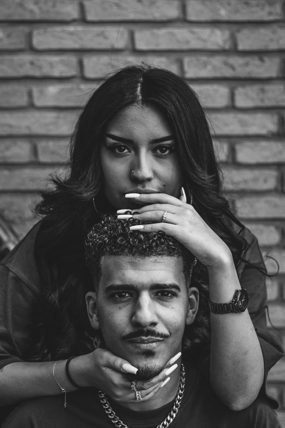 Black and white portrait of a couple against a brick wall: she stands behind him with her hands under his chin