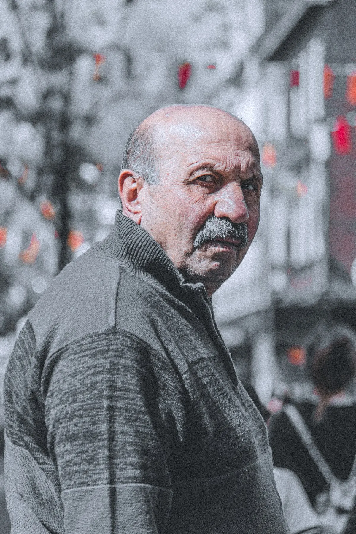 Portrait of an older man with a moustache and grey sweater looking over his shoulder, the street behind him in black and white