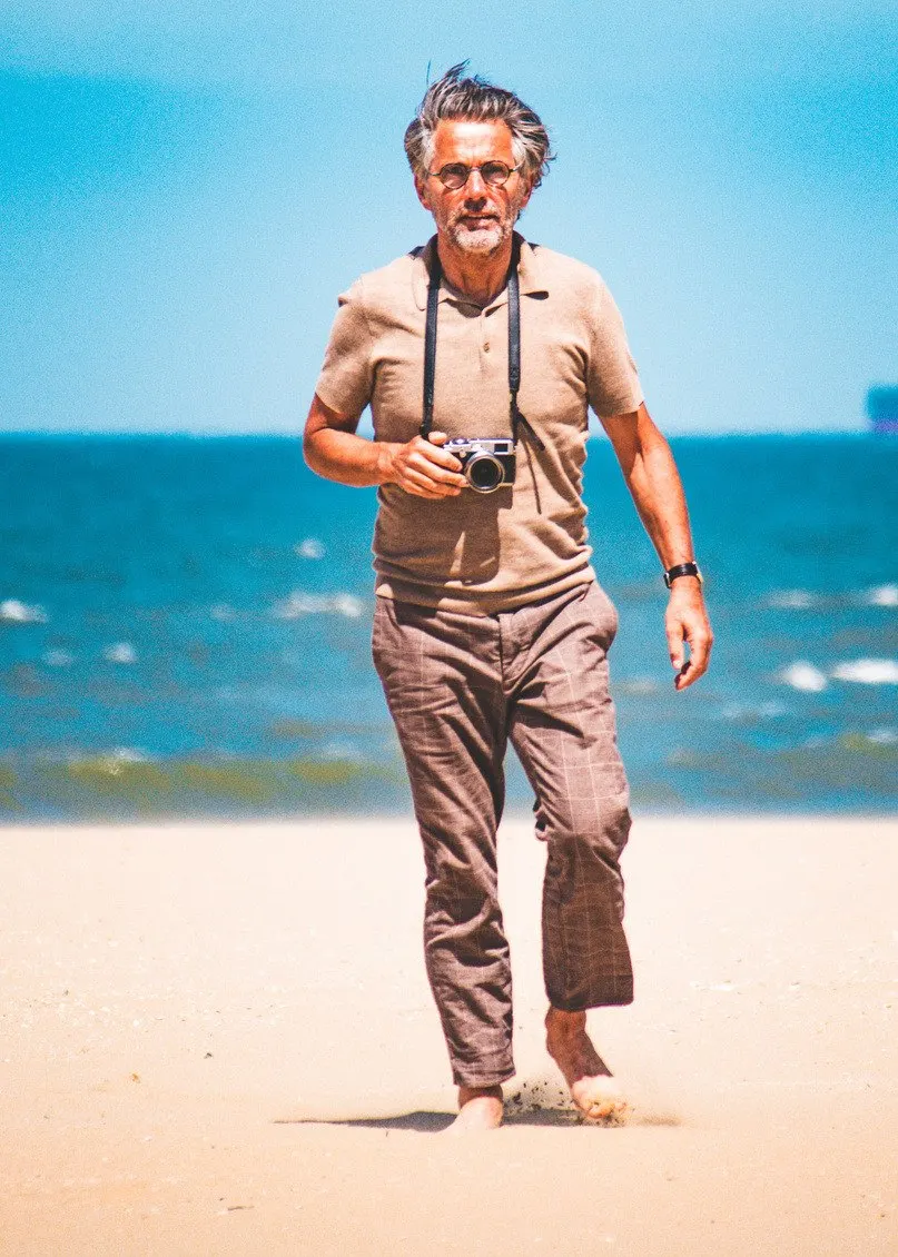 Grey-haired man with a camera around his neck walking barefoot on the beach, the sea behind him