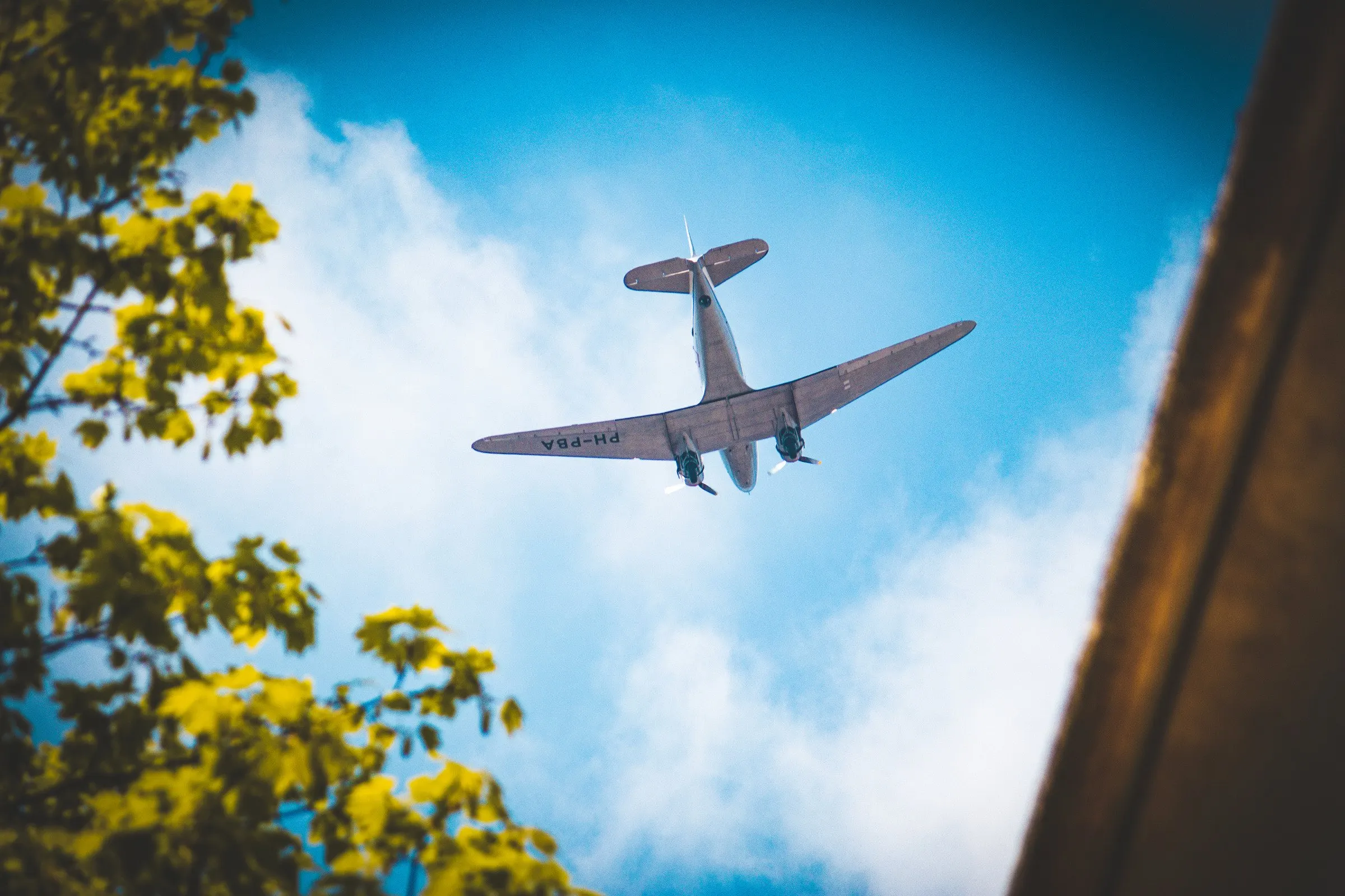 Propeller plane flying low overhead, seen between leaves and a building against a blue sky