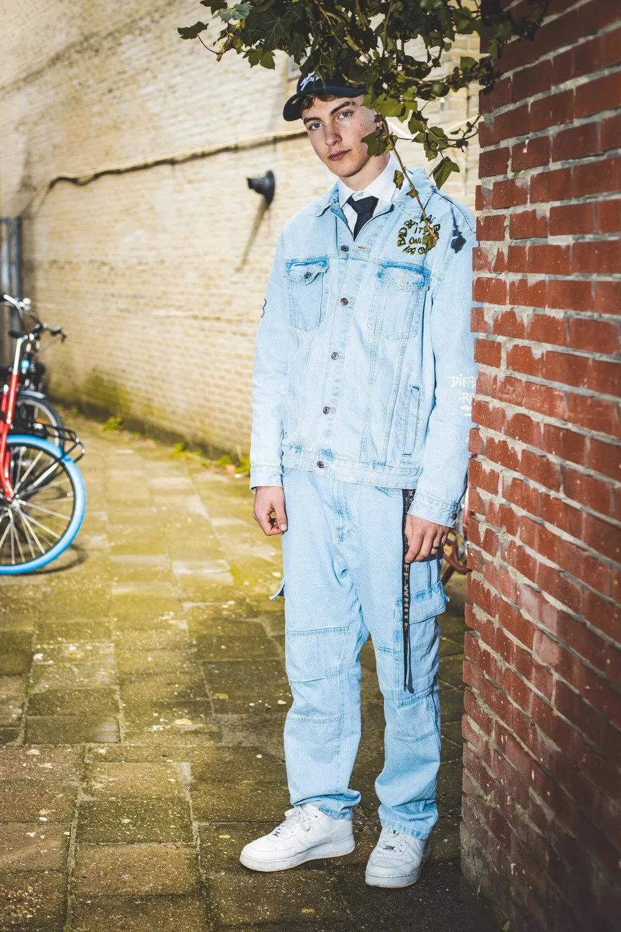 Young man in a light blue denim suit and cap leaning against a brick wall in an alley