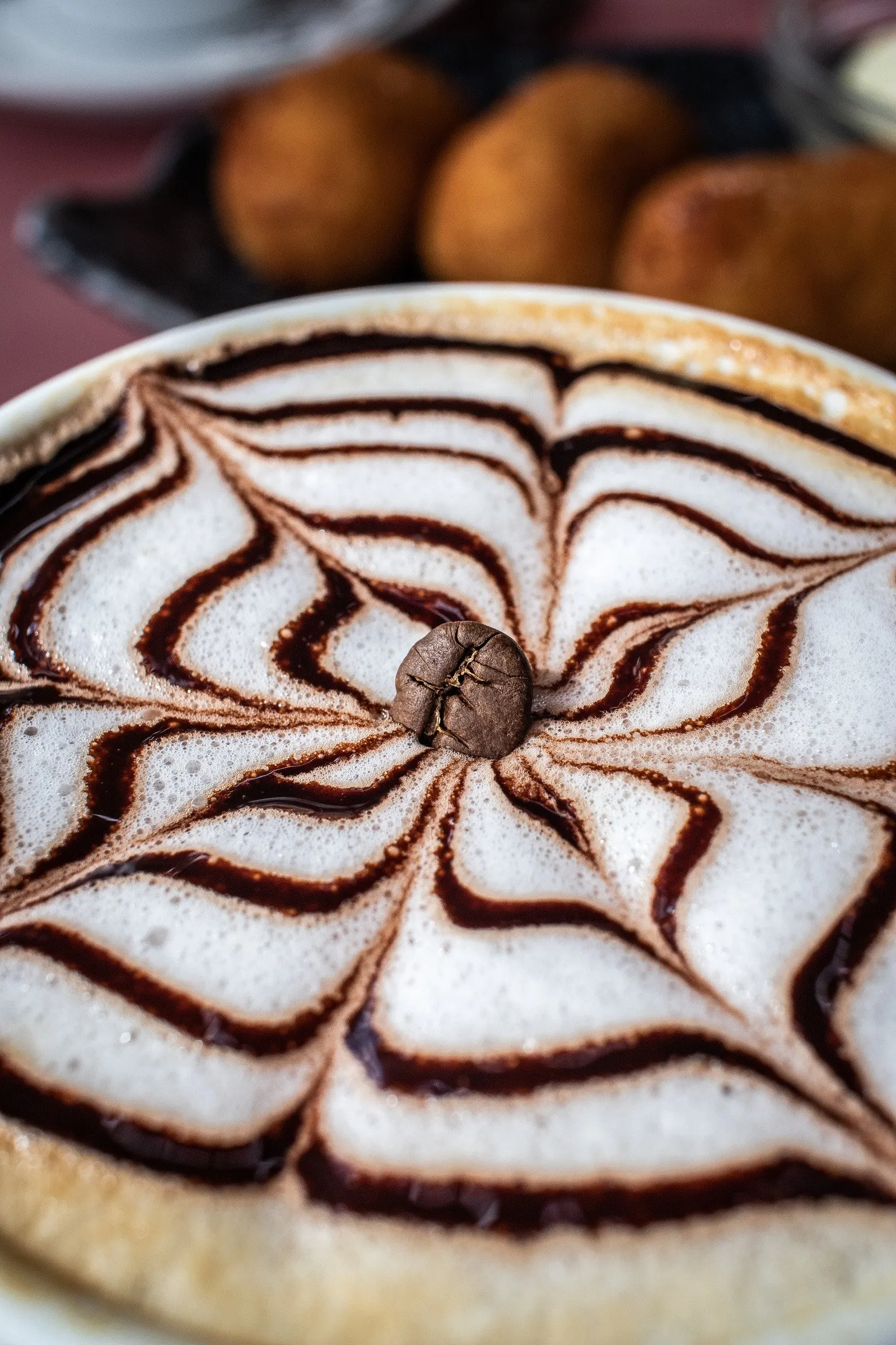 Cappuccino from above with a spiderweb of chocolate sauce, pastries in the background