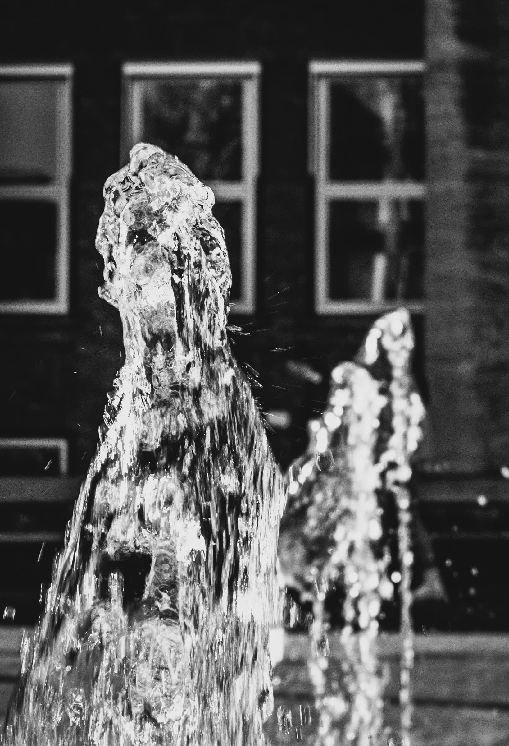 Black and white photo of water splashing up from a fountain in front of a building with windows