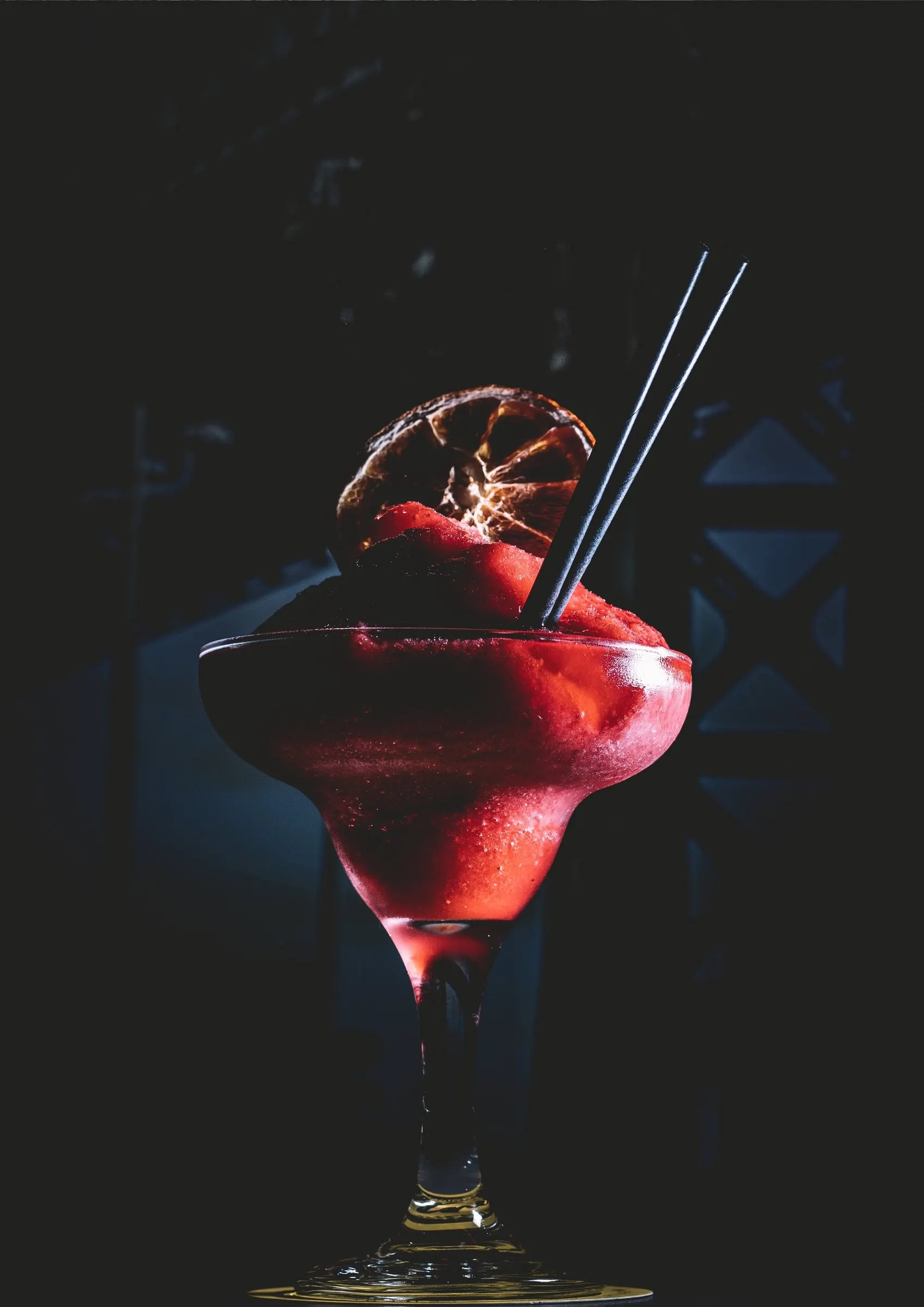 Red frozen cocktail with a dried orange slice and black straws against a dark background
