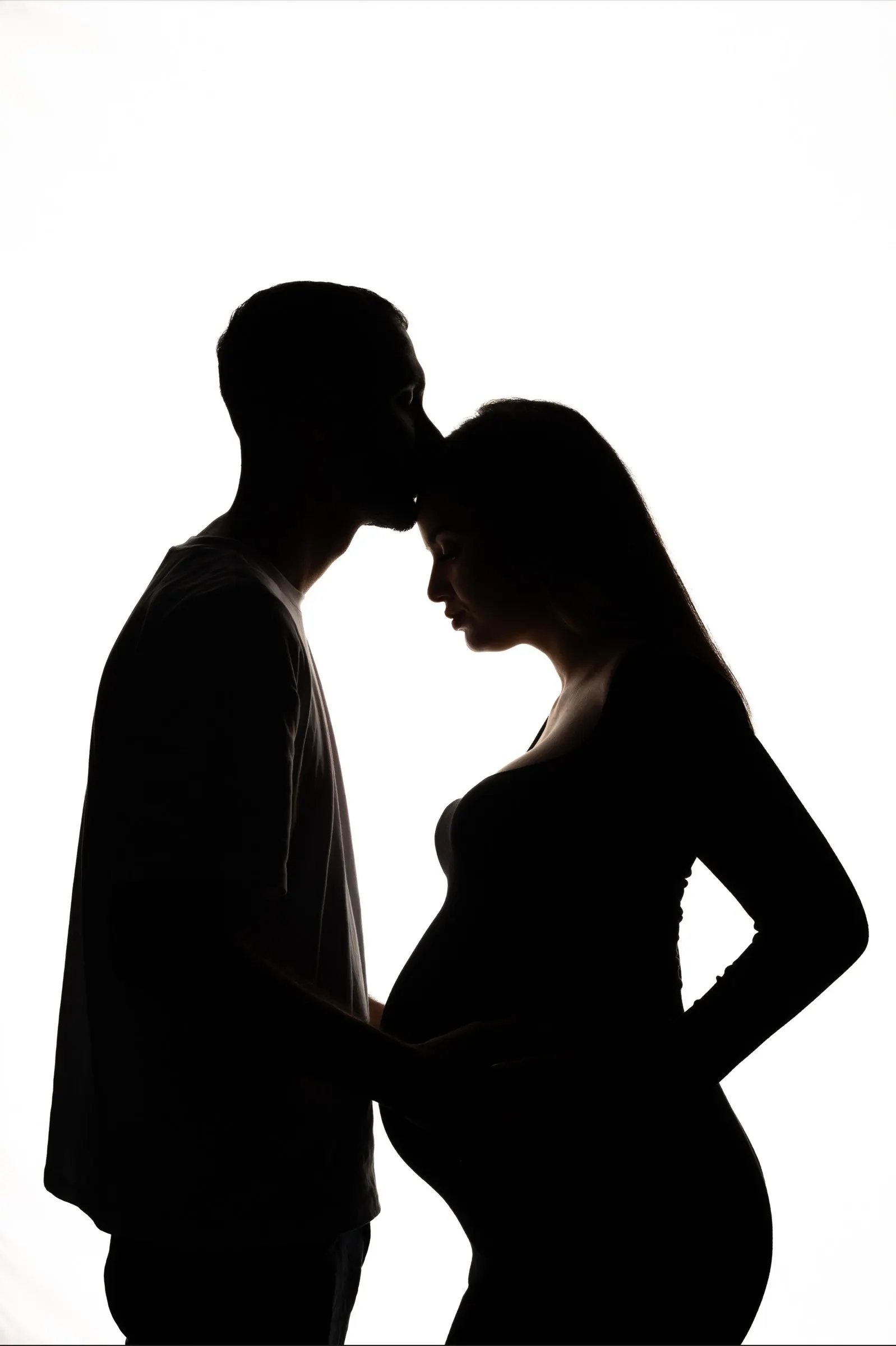 Silhouette of a couple against a white background: he kisses her forehead, she is pregnant